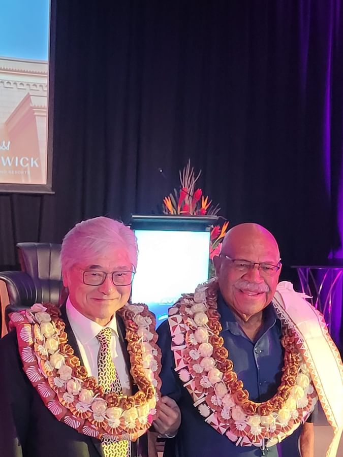 Two executives wearing traditional floral leis stand together at a Warwick Hotels event celebrating company milestones.