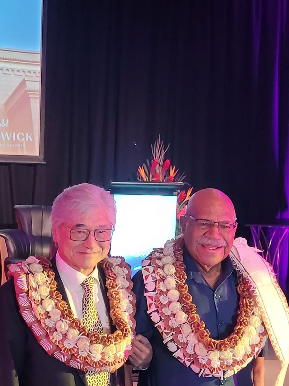 Two executives wearing traditional floral leis stand together at a Warwick Hotels event celebrating company milestones.