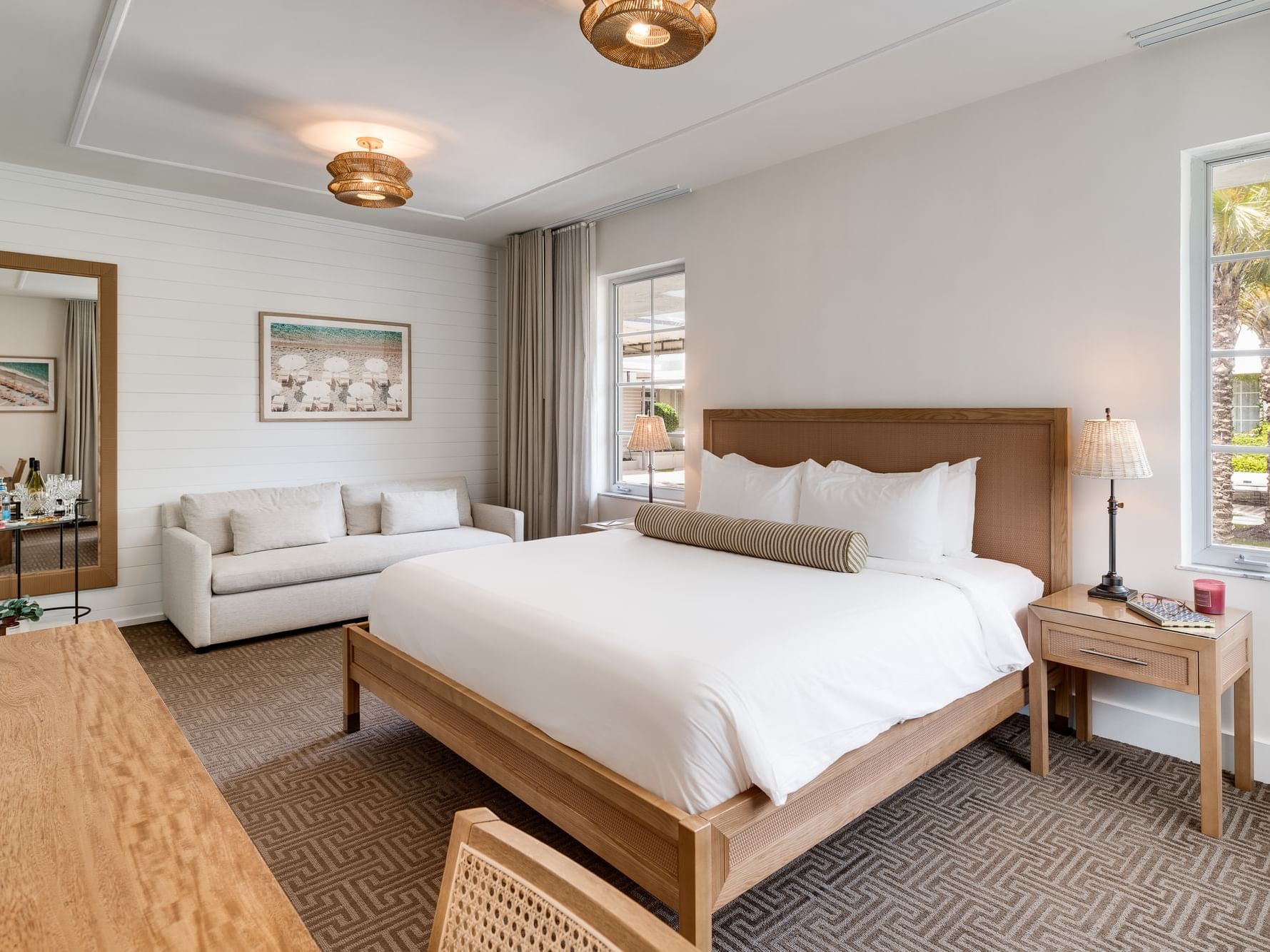 Spacious and stylish hotel room featuring a King bed with crisp white linens and a striped bolster pillow, a cozy white sofa, and large windows offering views of palm trees and the park courtyard. The room includes warm wood furnishings, a full-length mir