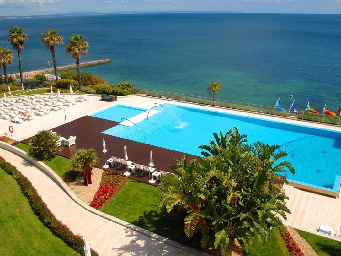 Aerial view of a swimming pool in Cascais, Lisbon, with surrounding greenery, beach chairs, and ocean in the background