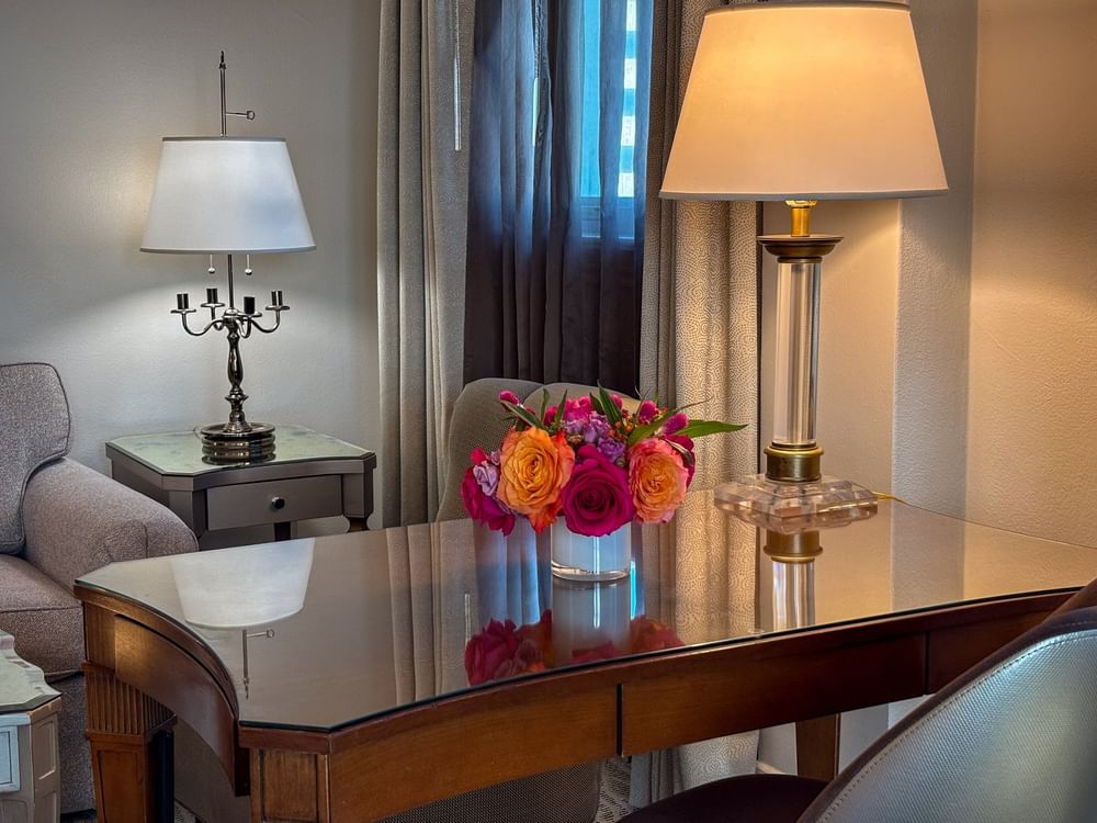 Wood desk with flowers by a mirrored side table under glowing lamps in Corner King at Warwick Melrose Dallas