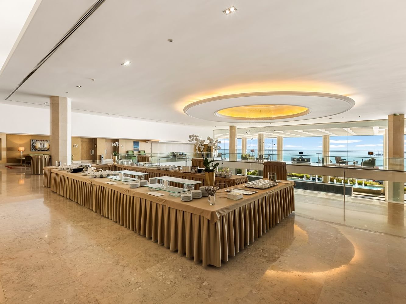 Galería de hotel con mesa larga y vista al mar, decorada con flores y cubiertos.