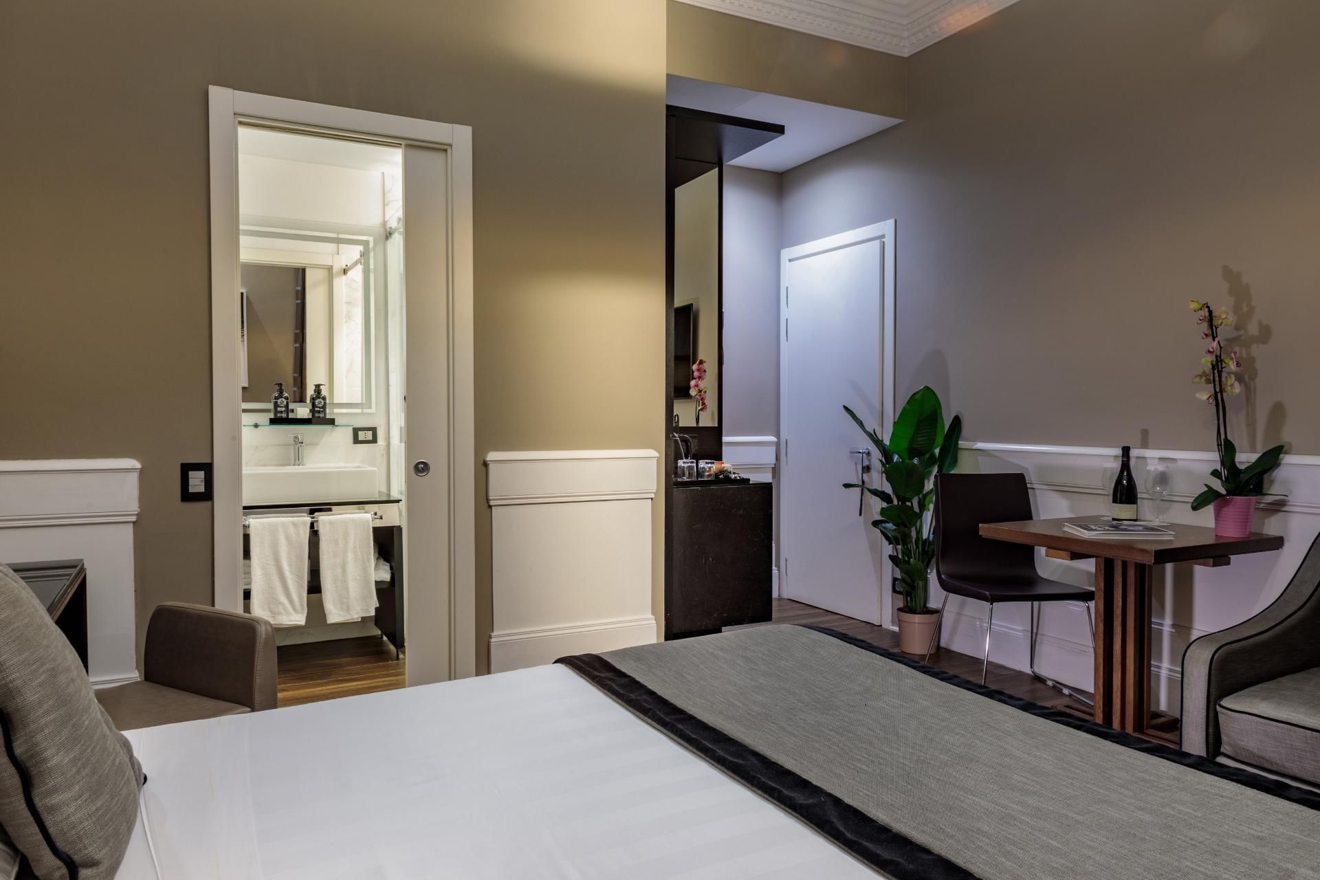 Stylish Deluxe Room at The Independent Suites with a cozy bed, a bathroom doorway, and a small table with plants and wine