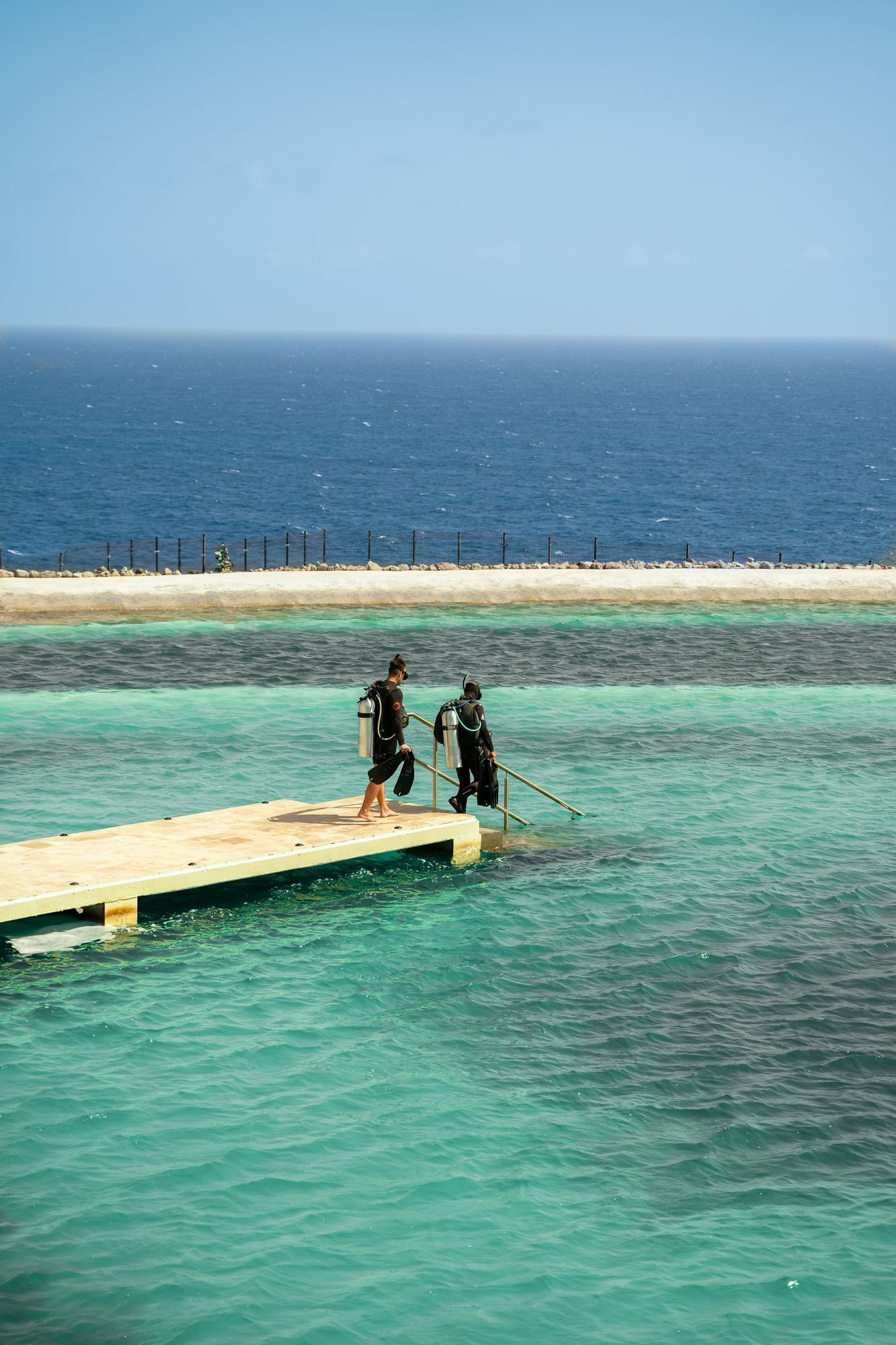 Twee duikers in wetsuits lopen een houten steiger op boven helder turquoise water nabij Golden Rock Resort