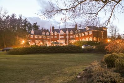 Evening view of a brick hotel building with warm lights and a green lawn under a cloudy evening sky at The Simsbury Inn