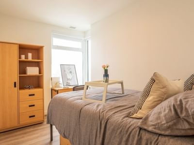 A bedroom bathed in light from floor to ceiling windows, and includes a bed, a desk, a desk chair, a dresser and a wardrobe.