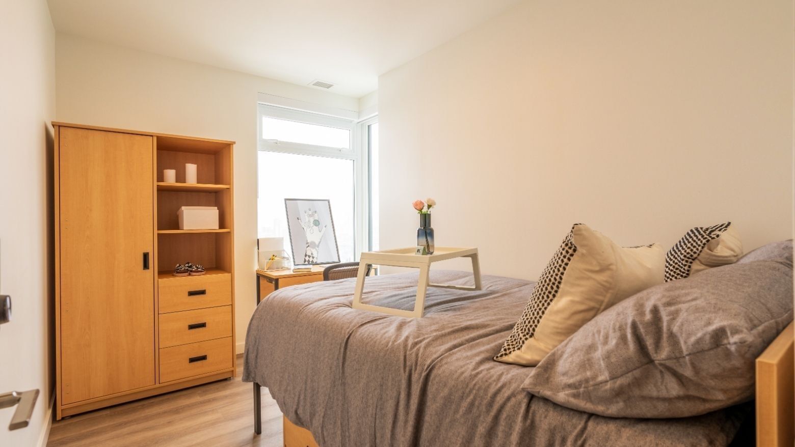 A bedroom bathed in light from floor to ceiling windows, and includes a bed, a desk, a desk chair, a dresser and a wardrobe.