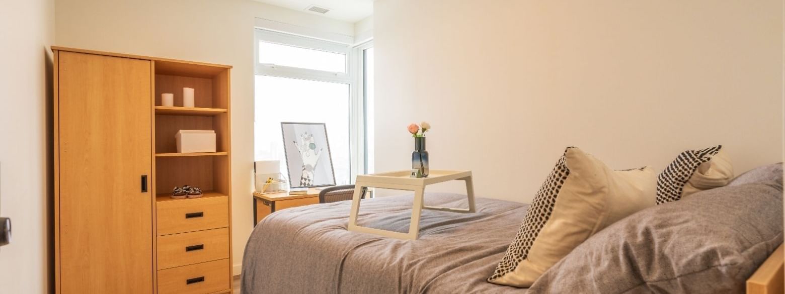 A bright bedroom in a shared apartment that includes a bed, under bed storage, desk, desk chair, dresser, and a wardrobe.