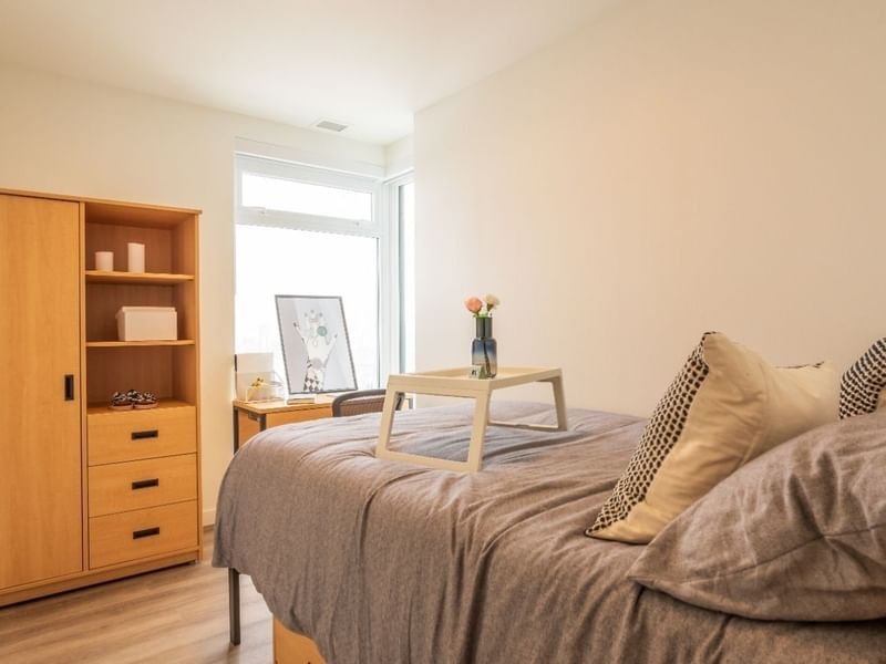 A bright bedroom in a shared apartment that includes a bed, under bed storage, desk, desk chair, dresser, and a wardrobe.