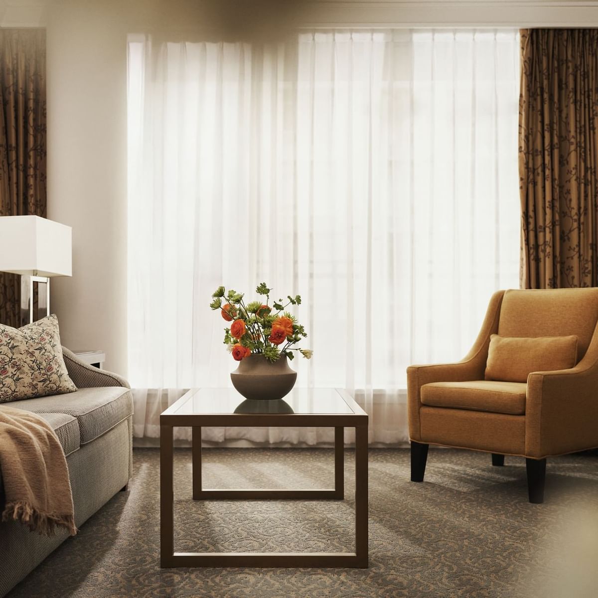 A living room with a couch, chair, table, and flower vase, illuminated by two lamps and curtains.