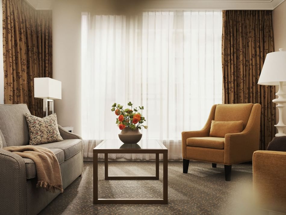 A living room with a couch, chair, table, and flower vase, illuminated by two lamps and curtains.