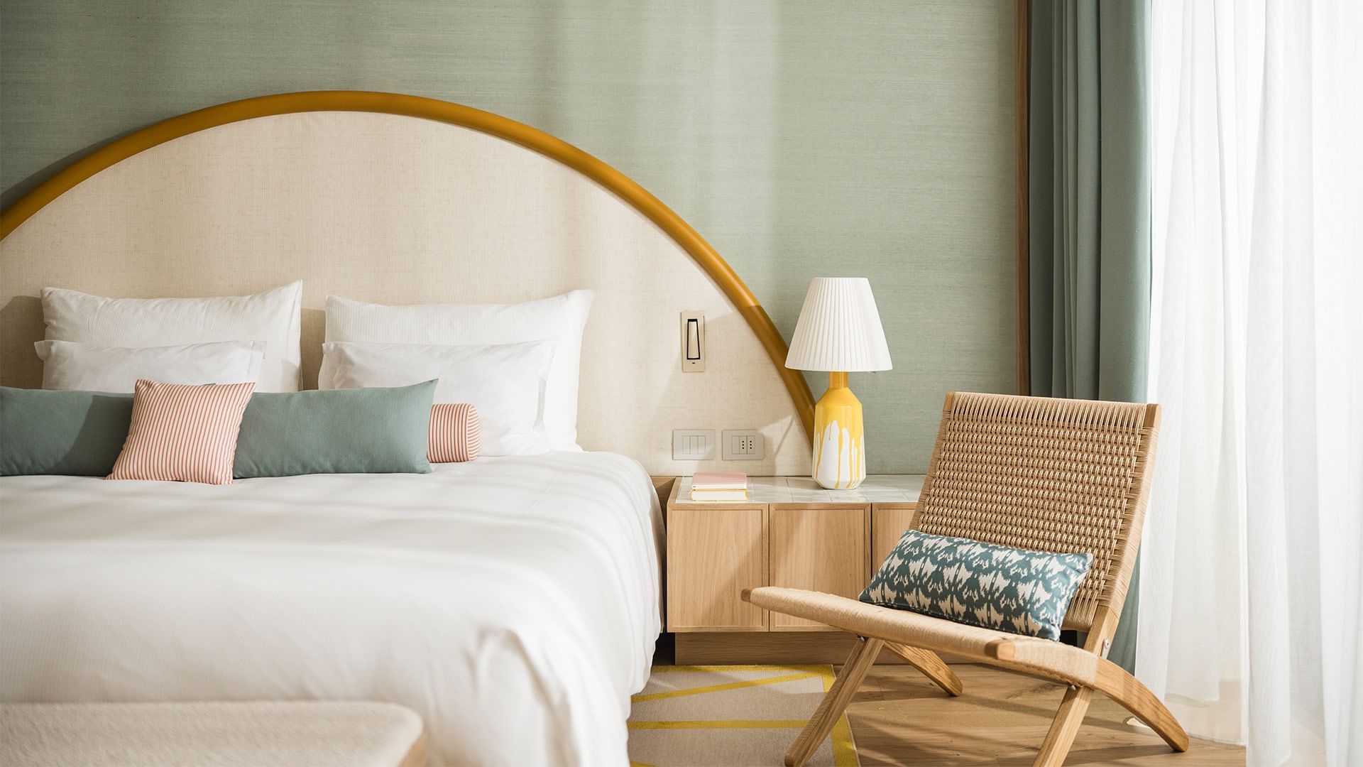 Bedroom with arched headboard, white bed, wicker chair, and nightstand with lamp and curtain in the background.