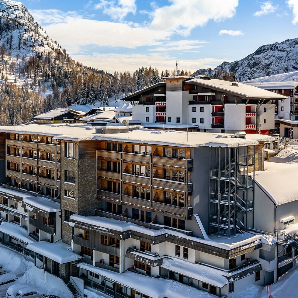 Complesso alberghiero innevato con montagne innevate sullo sfondo e un cielo blu.