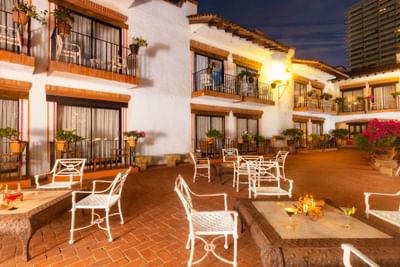 Evening view of a courtyard with tables, chairs, and buildings with balconies and potted plants.