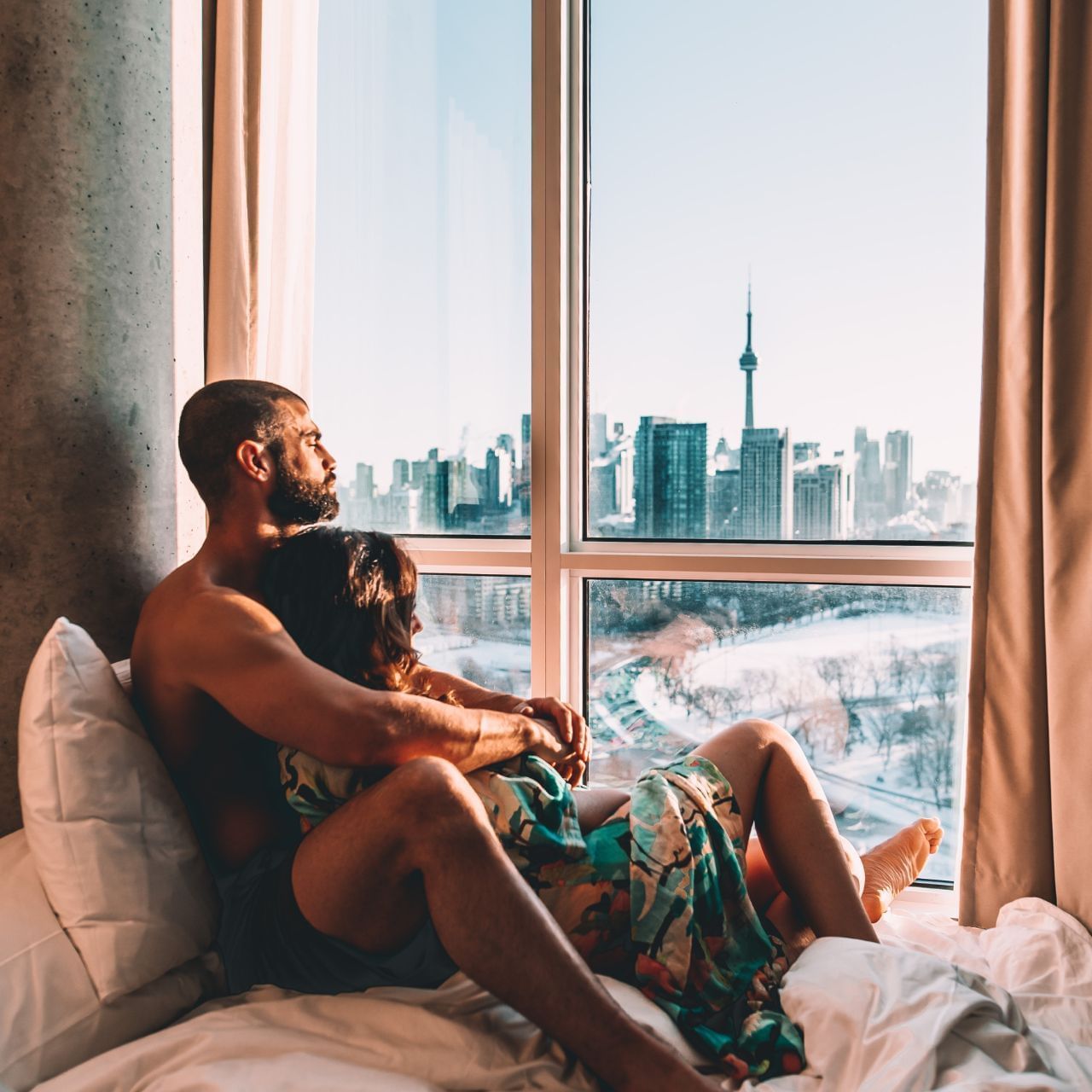 Couple relaxing on a bed facing expansive windows with panoramic skyline views of the city at Hotel X Toronto