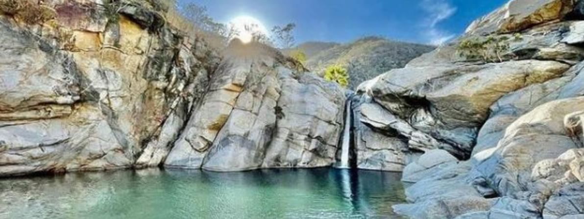Cascada cayendo en poza de agua turquesa rodeada de formaciones rocosas grises bajo cielo azul despejado.