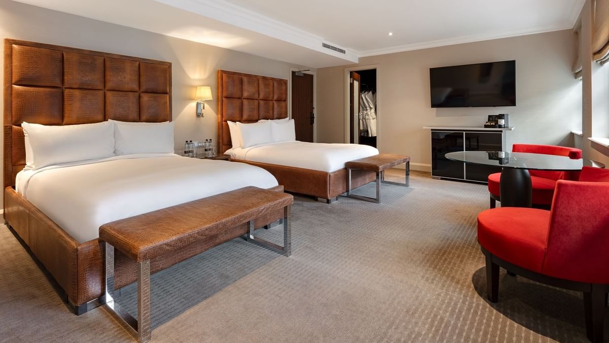 Spacious Family Room at The May Fair Hotel featuring two double beds, red chairs, a glass table, and a large TV