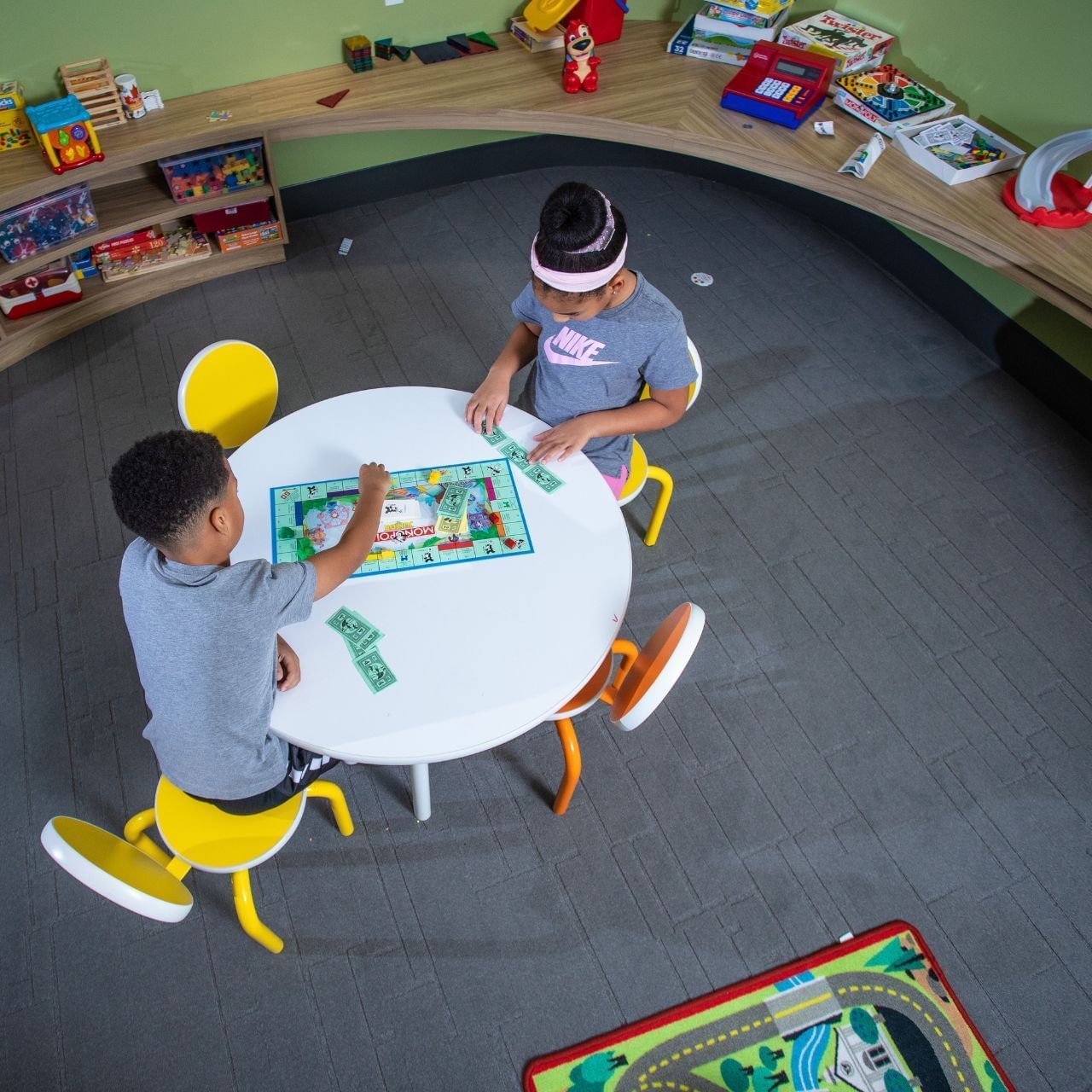 Two children playing Monopoly at a small round table in a playroom with toys and games.