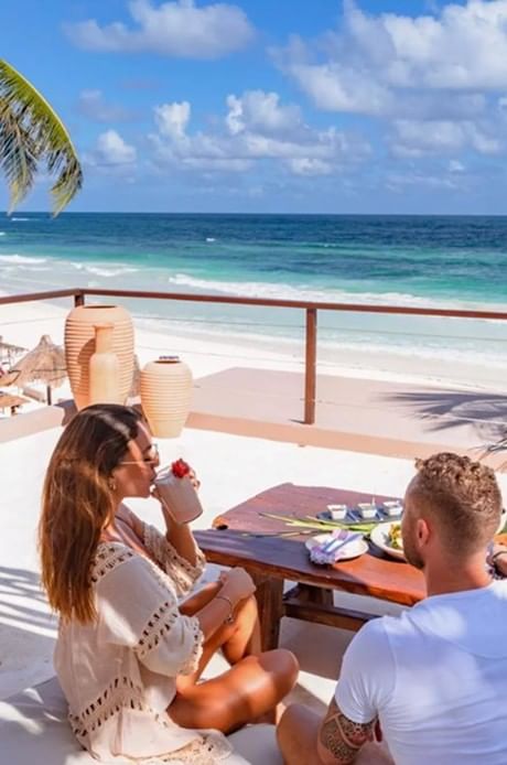 A couple enjoys a sun-drenched meal on a rooftop terrace overlooking the white sand beach at La Zebra Hotel