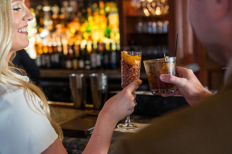 Two guests toasting cocktails by a wooden bar under a warm glow in the lounge at Warwick Melrose Dallas