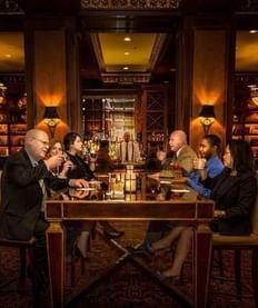 Group of guests enjoying drinks and conversation at a table inside a warmly lit lounge with bookshelves in The Library Bar