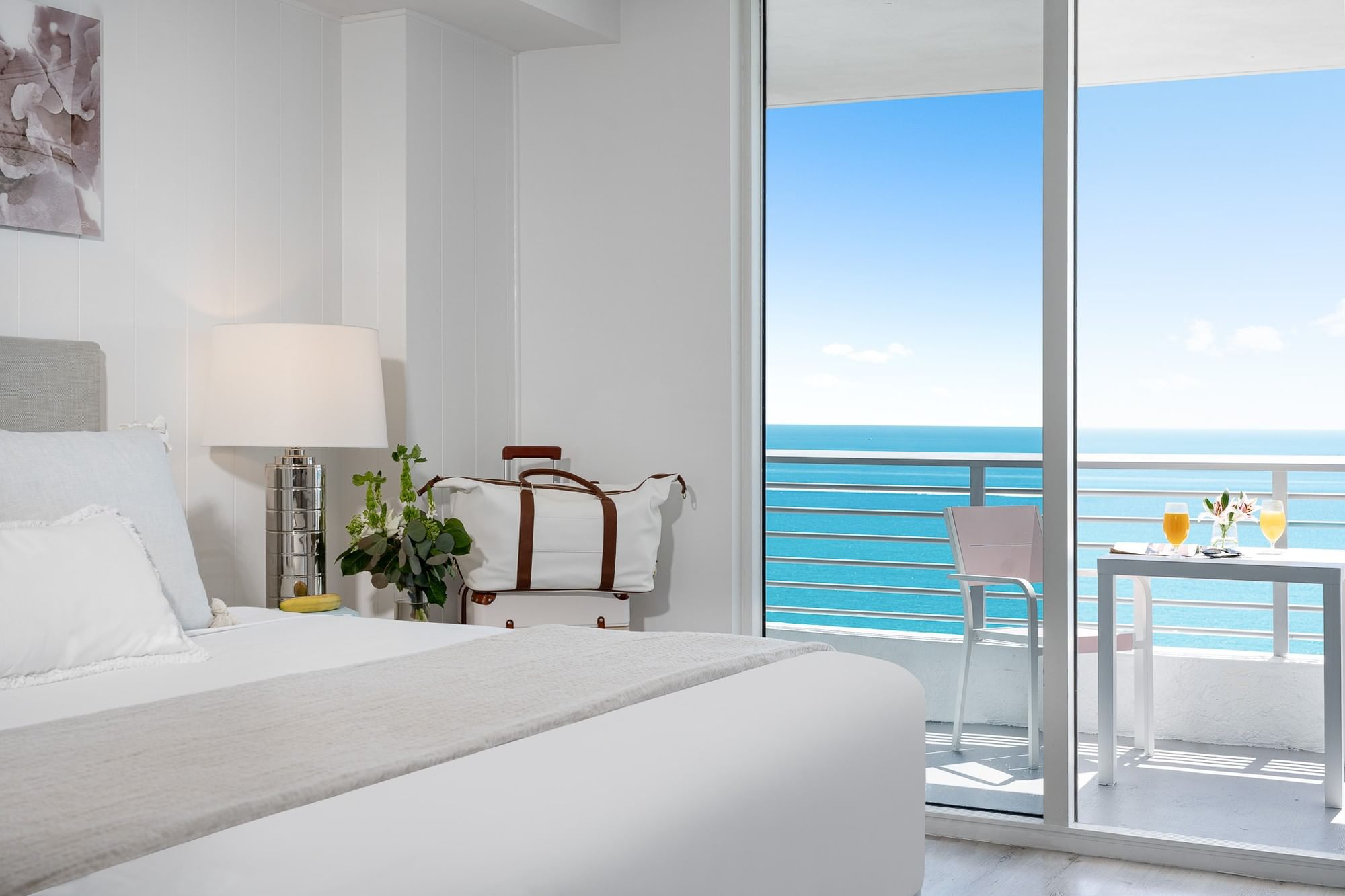 A white bedroom with a bed, lamp, and balcony overlooking the ocean.