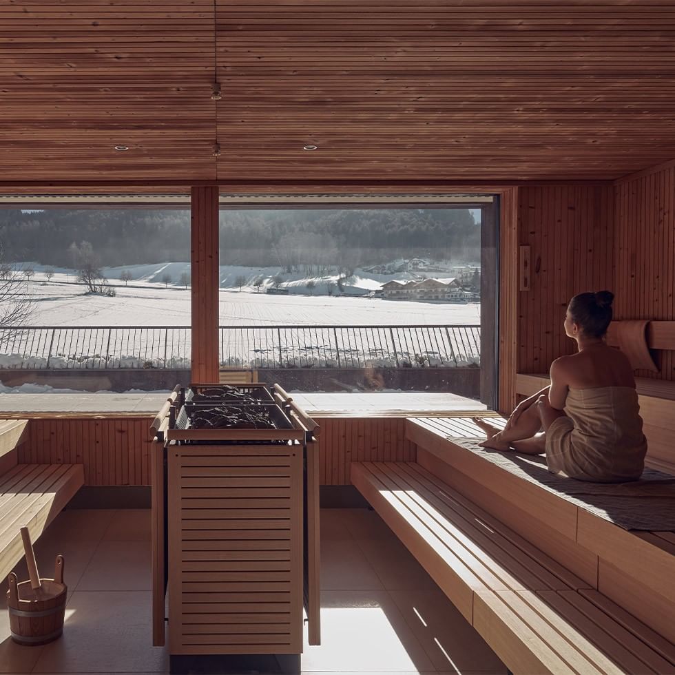 Donna in sauna in legno con vista su paesaggio innevato e prati, atmosfera rilassante.