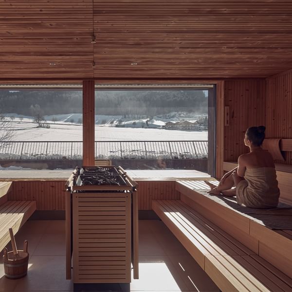 Woman in a towel sitting in a sauna with a large window showing a snowy landscape.