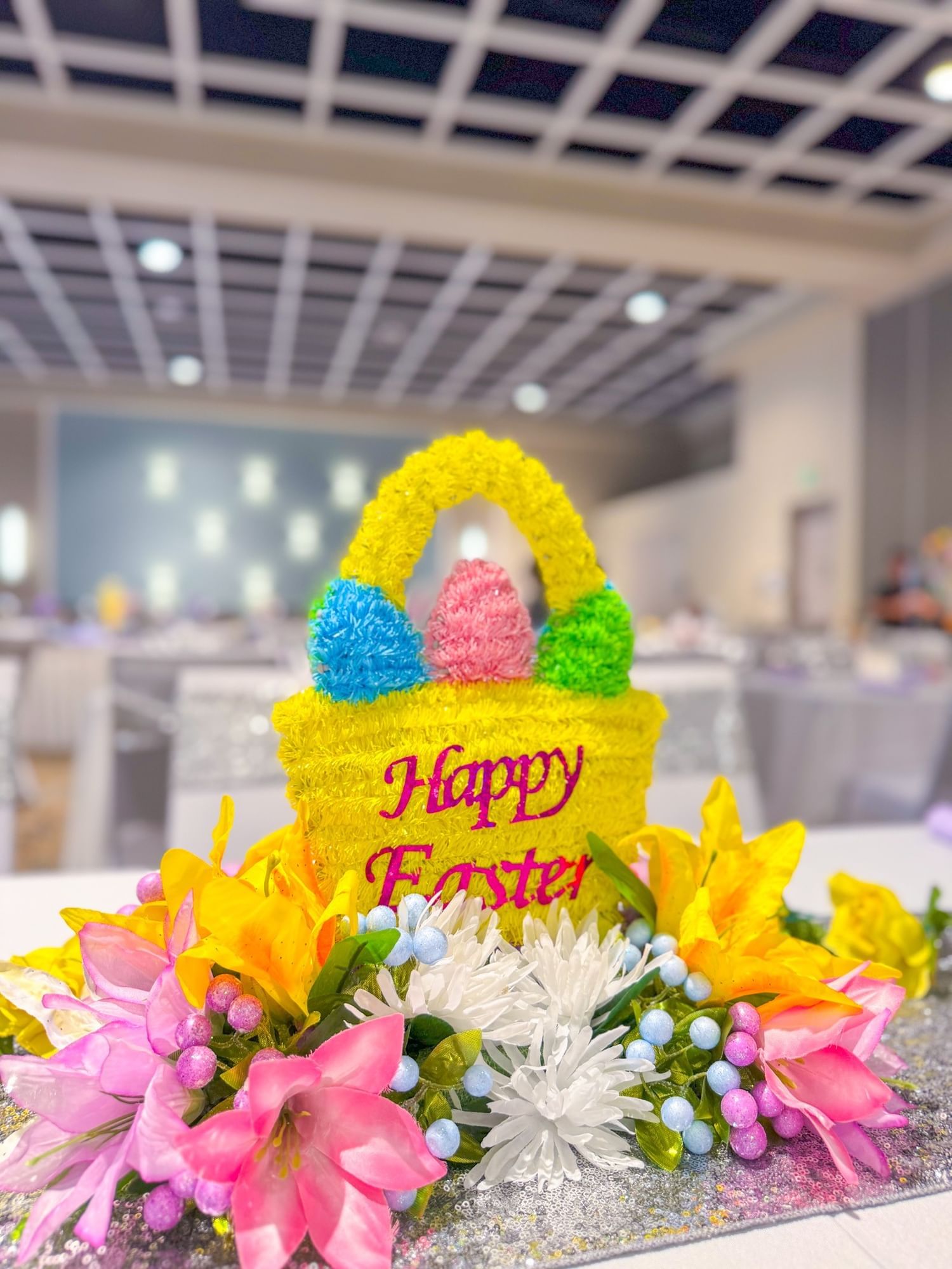 A large banquet room with a close-up on a centerpiece with a yellow basket that reads “Happy Easter” surrounded by pink, purple, yellow, and white flowers.  