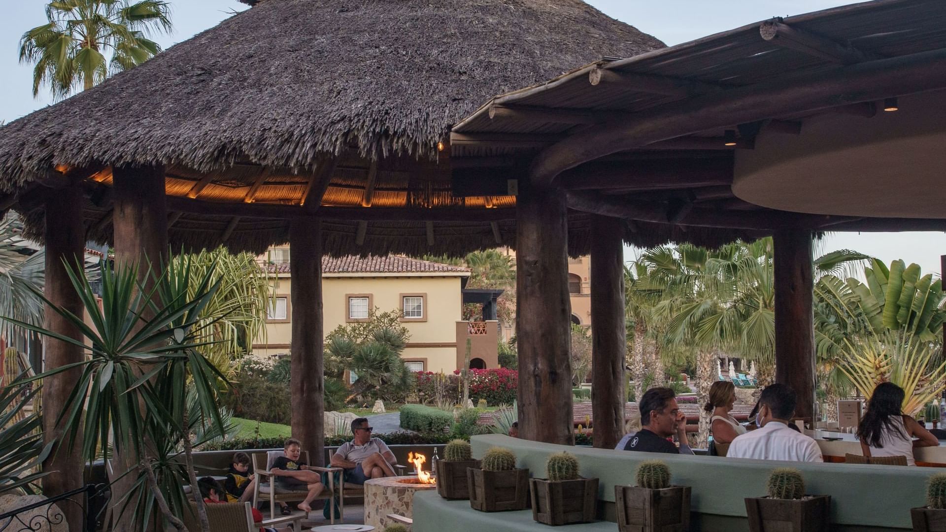 Clientes Relajándose junto a una fogata bajo un gran bar con techo de paja en el restaurante Pitahayas de Hacienda Del Mar Los Cabos