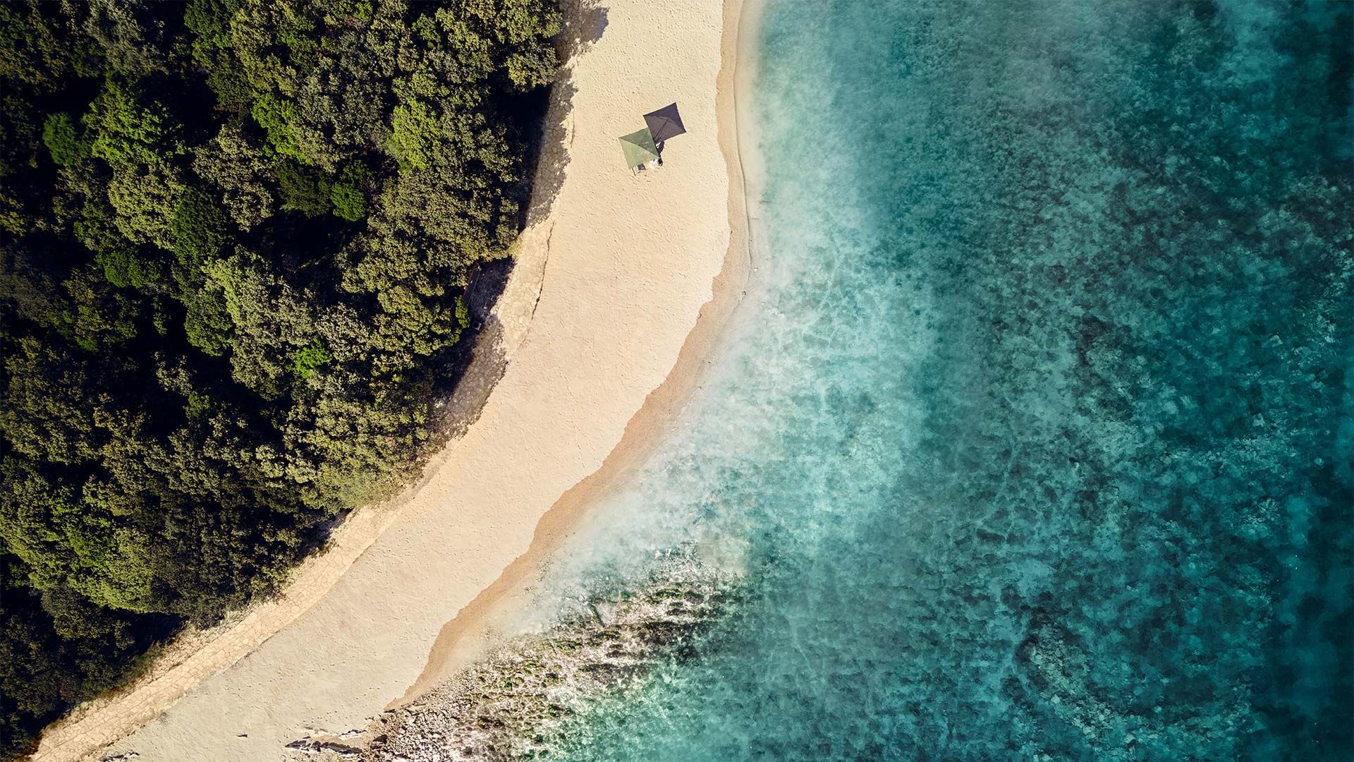 Zračni pogled na pješčanu plažu i bistro plavo more s zelenim šumovitim područjem.