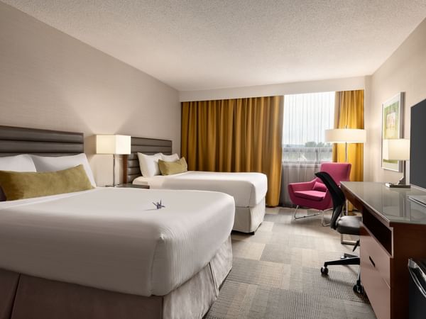 Two queen beds, desk with TV, chair, and window with curtains in hotel room.