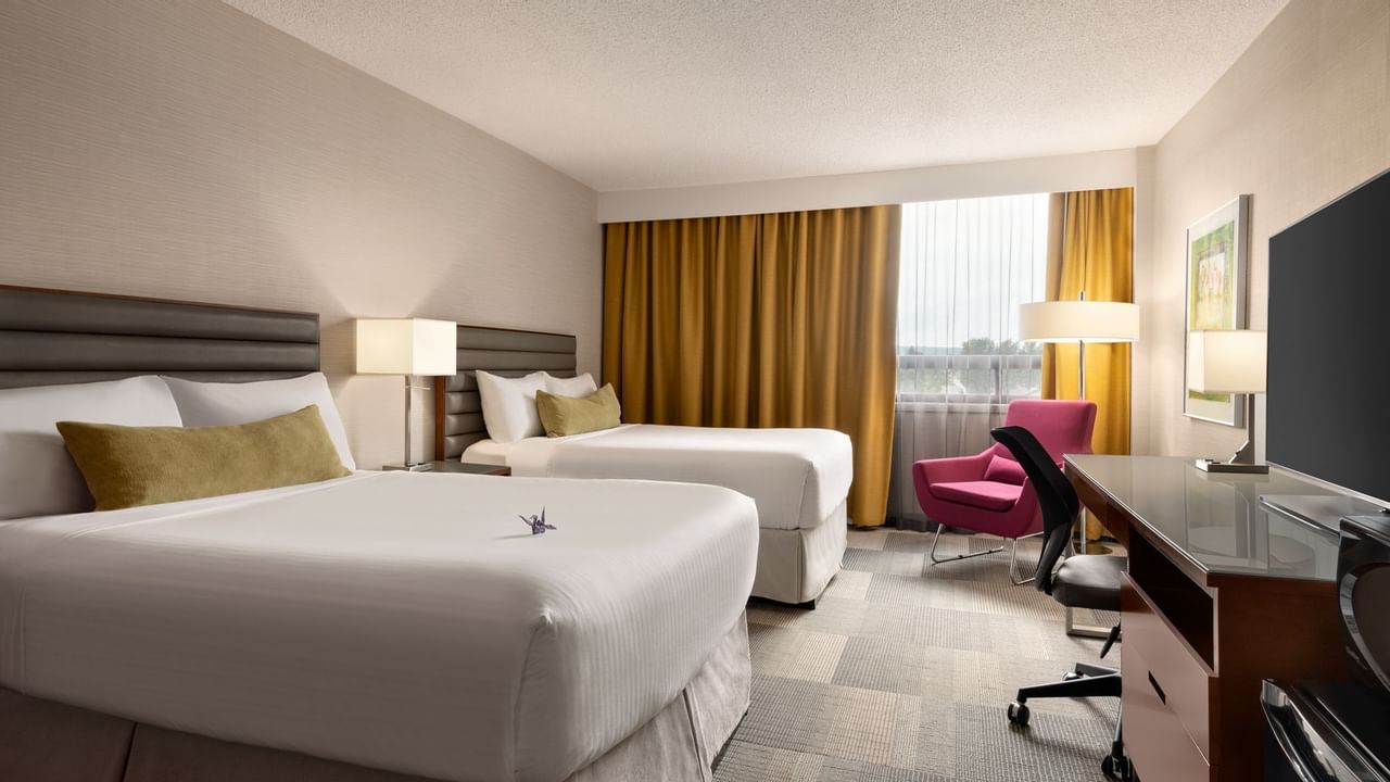 Two queen beds, desk with TV, chair, and window with curtains in hotel room.