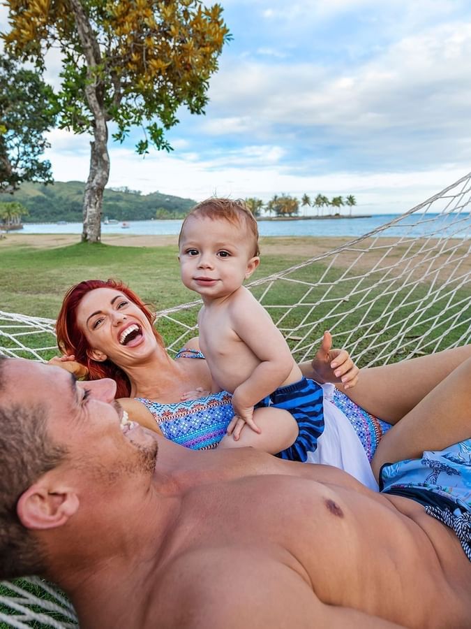 Laughing family relaxing in a large rope hammock on a lush green lawn by the ocean at The Naviti Resort - Fiji