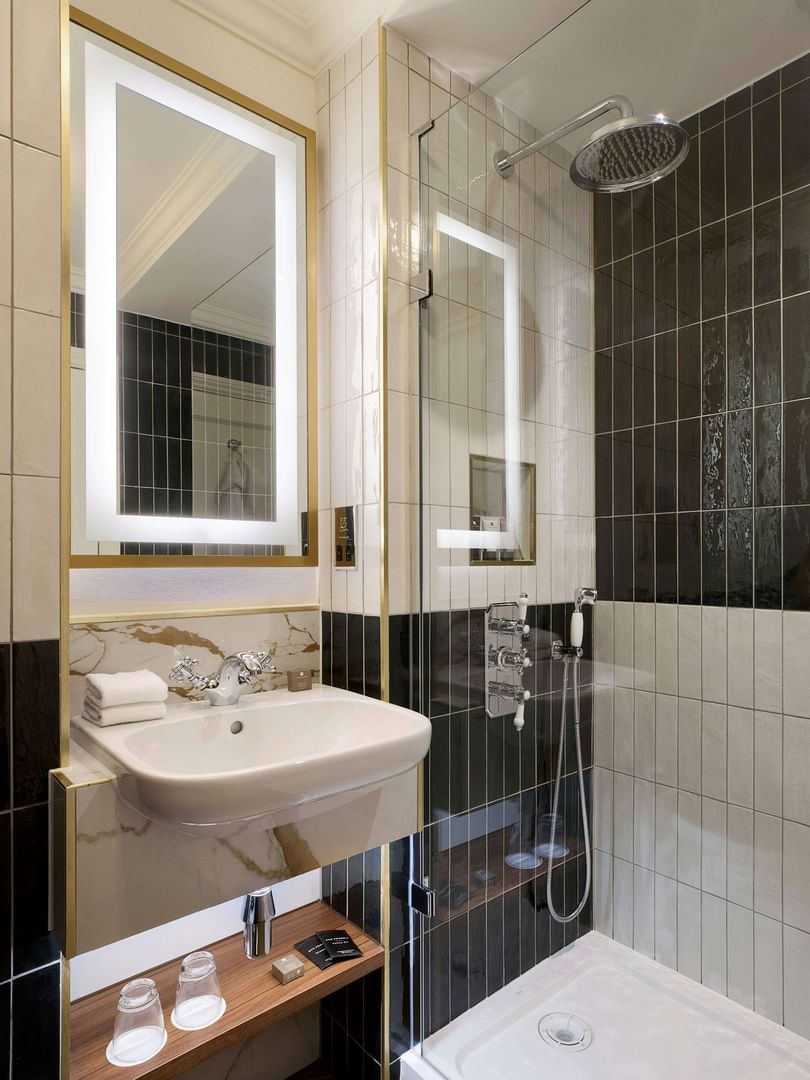 Modern bathroom with illuminated mirror, marble sink, glass shower, and black and white tiled walls at The Capital Hotel.