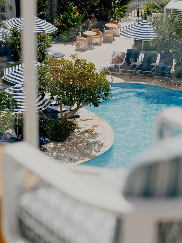 Curved pool with turquoise water by striped umbrellas and sun loungers under a leafy tree near guests at Elysium Hotels