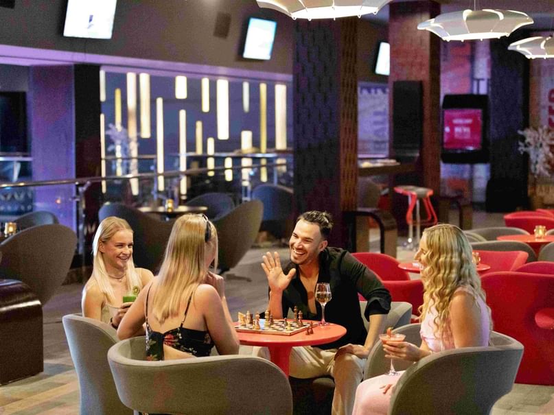 Four people sit at a table in Sports Bar , one playing chess while others chat and drink.