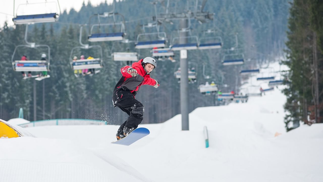 Snowboarder in red jacket and black pants rides down snowy slope past ski lift with passengers.