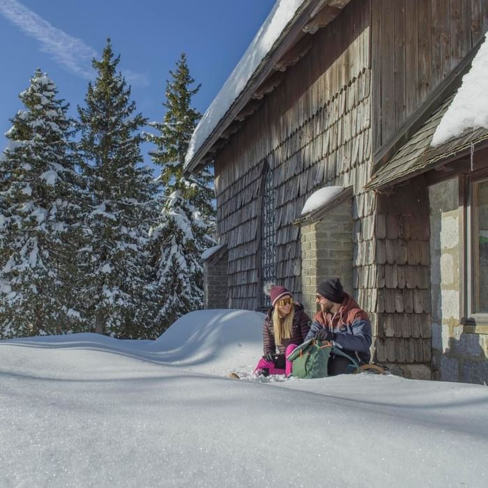 Coppia seduta sulla neve vicino a una casa in legno con tetto in legno e alberi innevati.