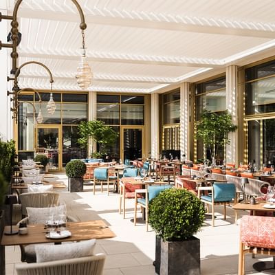 An elegant open-air restaurant with wooden tables, colorful chairs, and decorative plants under a white canopy.