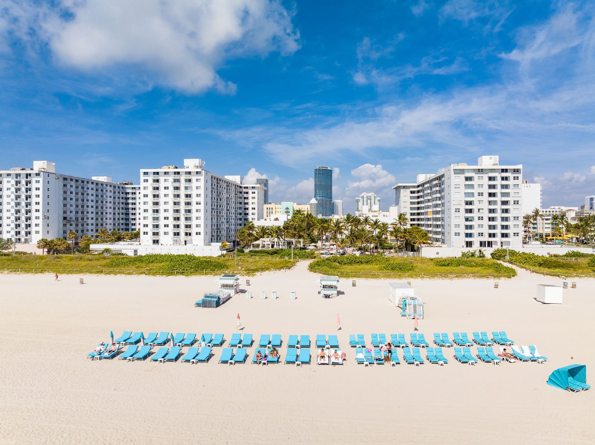A wide-angle view of a beach with lounge chairs, buildings, and palm trees in the background.