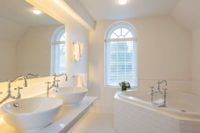 Clean bathroom of Jacuzzi Suite with two sinks and a large tub under a bright arching window at The Exeter Inn
