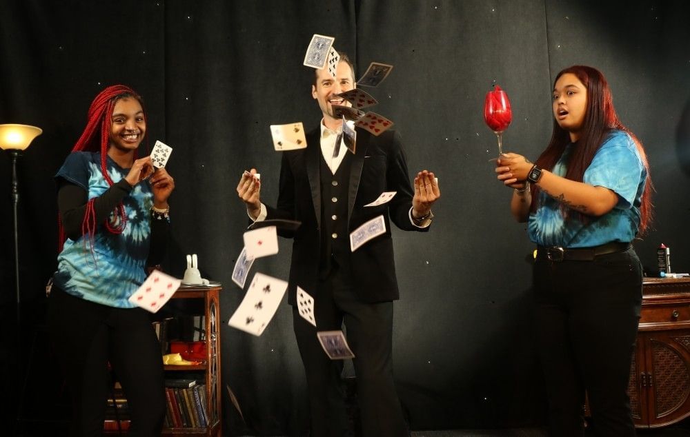 A man in a suit tosses a deck of cards into the air with two women in blue tie dye shirts on either side of him, smiling.
