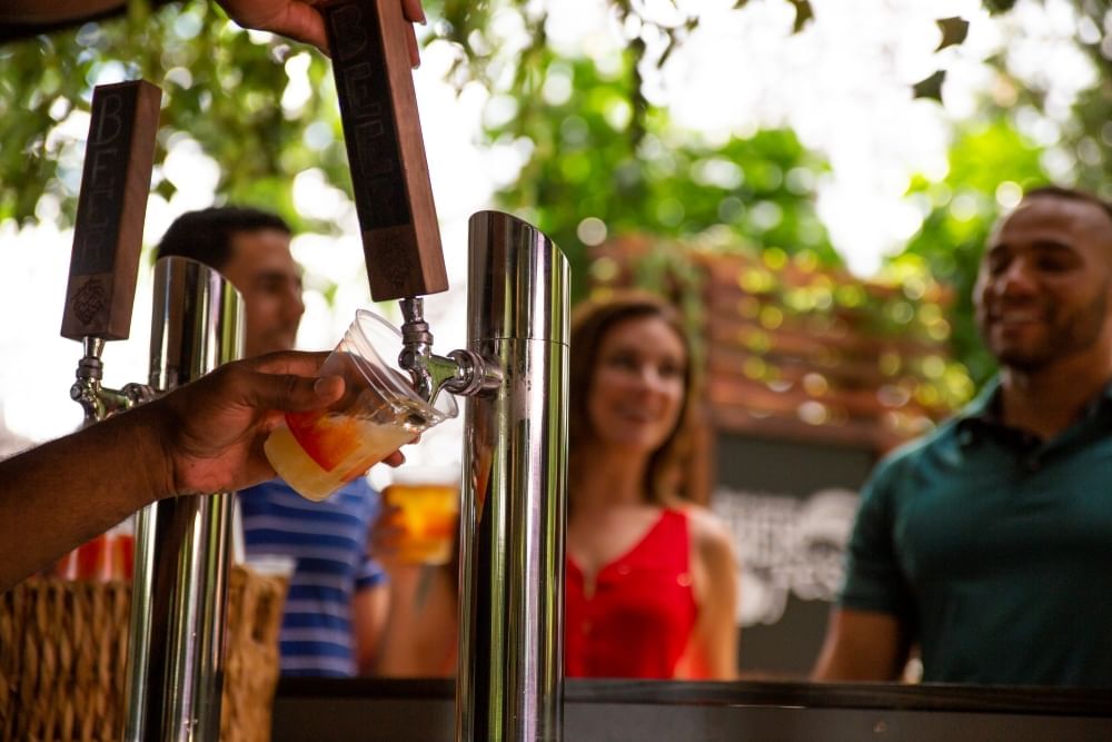 A hand pours a beer from a tap outdoors with three out-of-focus guests in the background.
