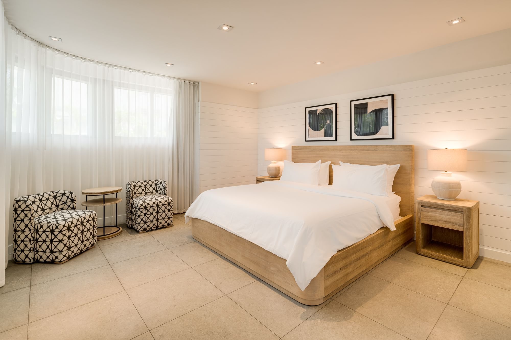 A wide view of a hotel room with a king-sized bed, two nightstands with lamps, two armchairs, and artwork on the wall.