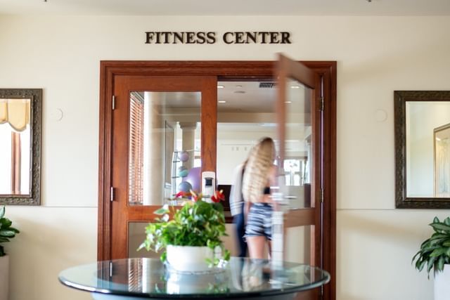 A doorway to the fitness center with a sign above it, flanked by mirrors and a plant on a table.