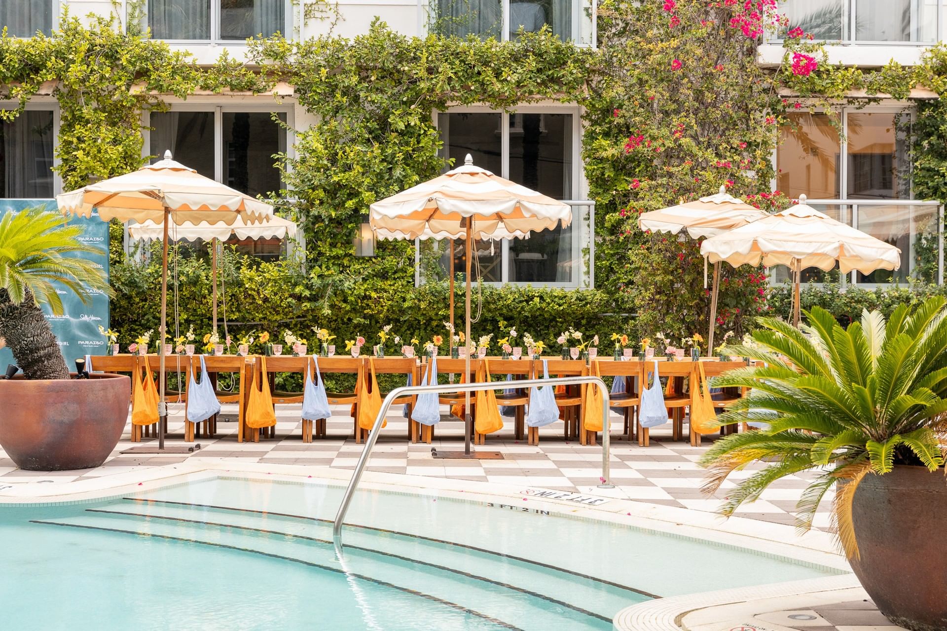 A poolside area with tables, chairs, and umbrellas surrounded by lush greenery and a building in the background.