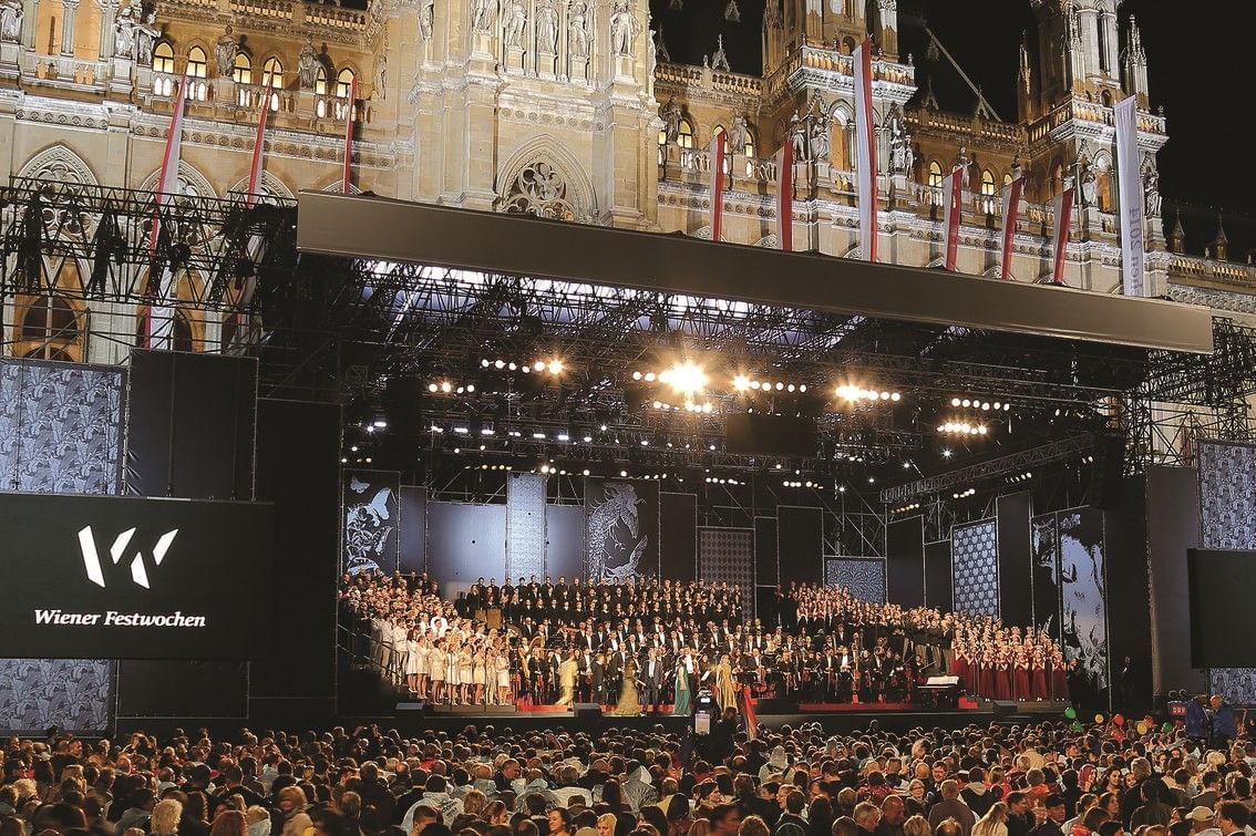 Performance of the Vienna Festival Weeks in front of Vienna City Hall featuring a large stage, orchestra, choir, and a large audience at Rathausplatz at night.