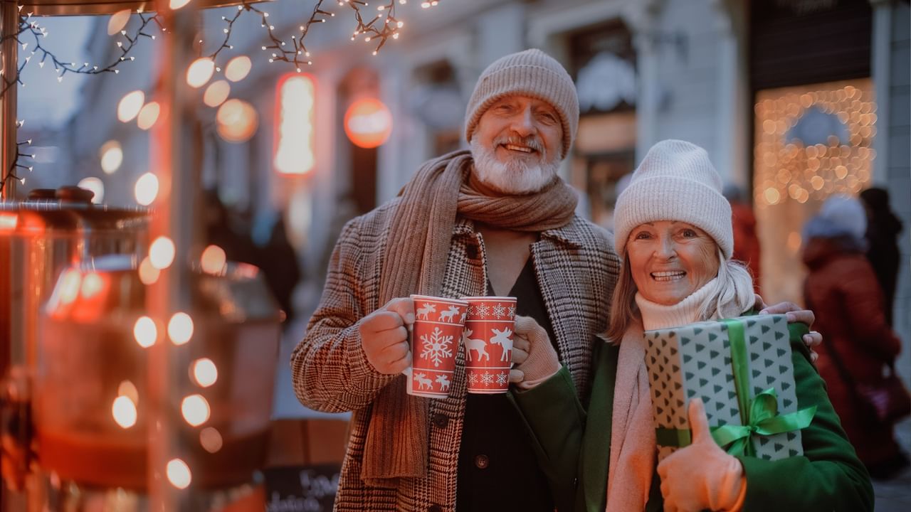 Elderly couple holding mugs and a gift, smiling, at a festive outdoor market.