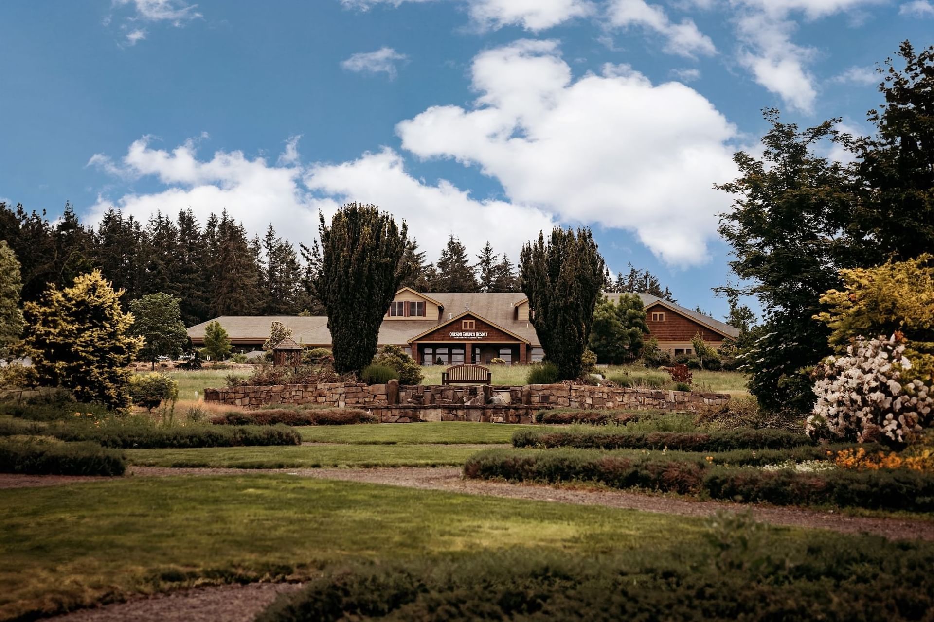 Lush garden with stone wall, trees, and bench in front of a large building under a partly cloudy sky.